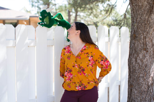 Kati of Hooked by Kati looking over her shoulder at a green crochet dragon sitting on a fence. 