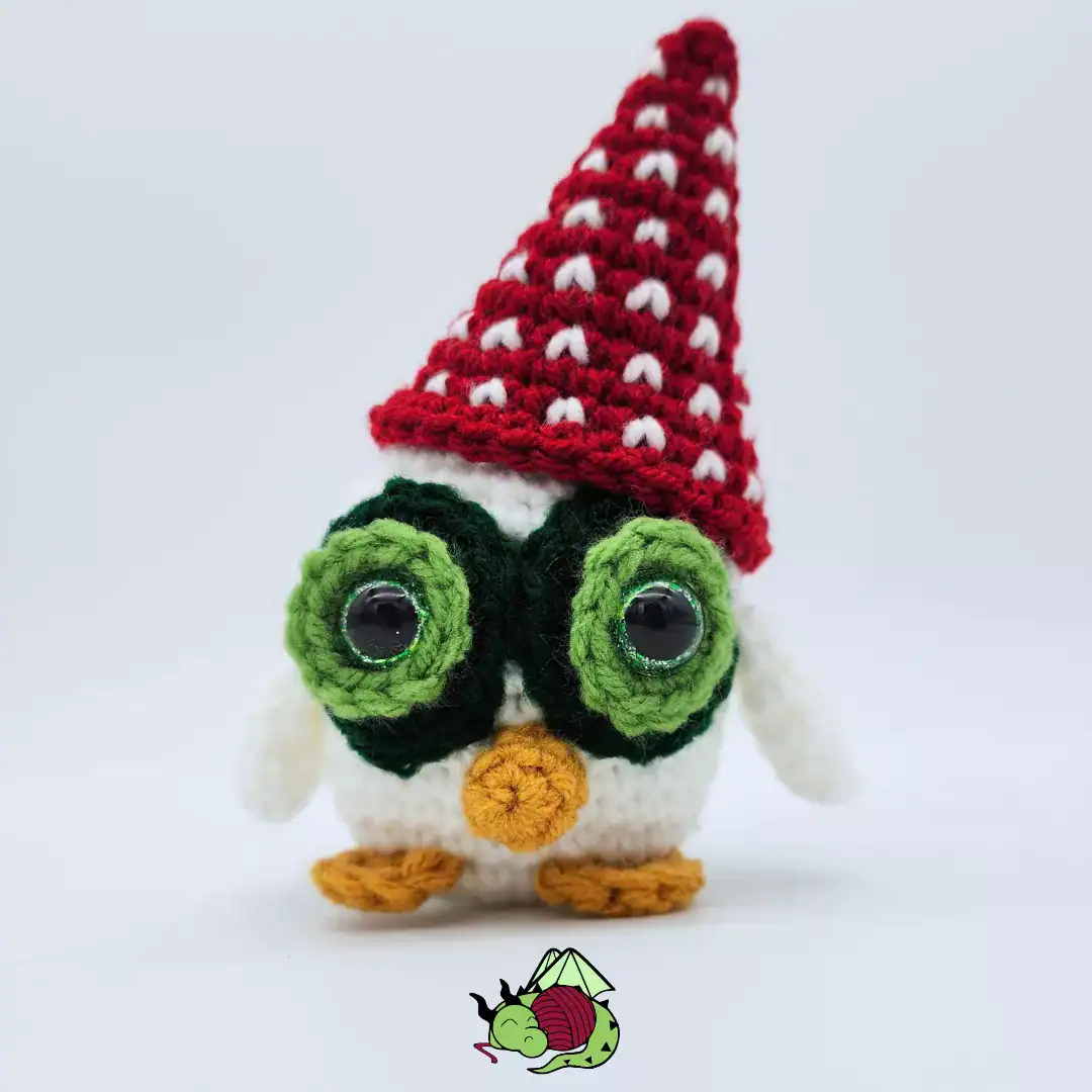 Little white crocheted owl with green eye circles wearing a crochet red pointed hat with white sts decorating it.
